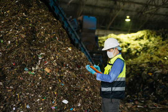 Quality inspector in hardhat and mask taking notes beside a large compost pile, evaluating organic waste management