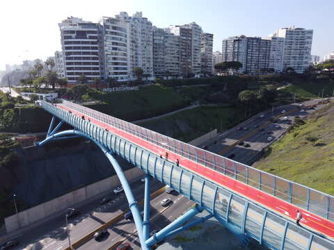 Aerial view of the modern Puente de La Paz pedestrian bridge with a red walkway spanning over a highway, surrounded by high-rise buildings and green cliffs in Miraflores, Lima Province, Peru.