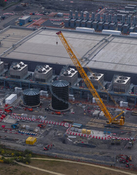 Aerial view of a hyperscale data center construction site featuring a large yellow crane, industrial cooling towers, and storage tanks in Belgium.