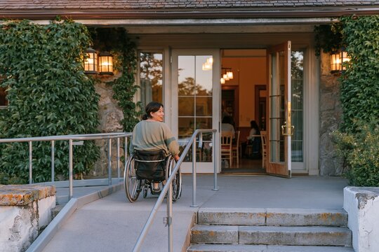 East Asian woman in a wheelchair using a modern access ramp to enter a historic stone building with glowing lanterns at dusk