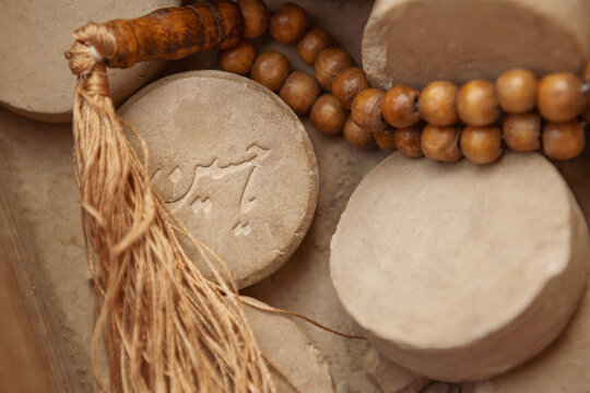 High-angle, close-up shot of Islamic clay prayer stones (Mohr or Turbah) alongside a wooden rosary (Tasbih). One prominent clay tablet features carved Arabic calligraphy reading "Ya Hussein".