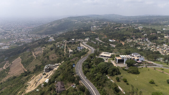 Aerial view of a winding road cutting through the lush green hills of Aburi with scattered buildings and a distant town view in Aburi, Eastern Region, Ghana.