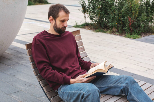 Bearded man with short brown hair reading open book while sitting on wooden bench outdoors, conveying education, self development, concentration, leisure, knowledge and thoughtful lifestyle