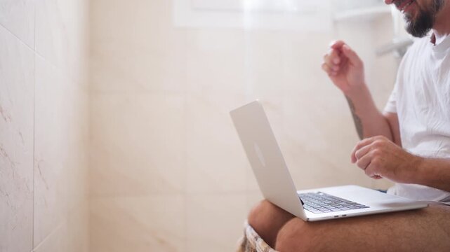 Excited bearded man using a laptop computer while sitting on the toilet, celebrating a successful deal or online win with a fist pump gesture, illustrating the concept of workaholism
