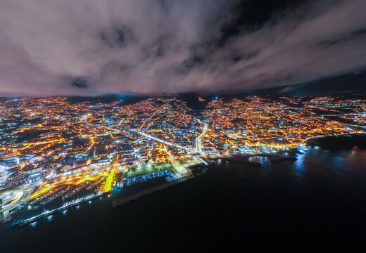 Aerial view of the illuminated cityscape of Funchal at night, featuring the harbor, coastal lights, and surrounding hills under a cloudy sky, Funchal, Madeira, Portugal.