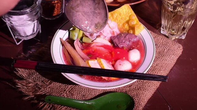 Close up of a spoon pouring vibrant red broth into a traditional Thai pink noodle soup bowl filled with fish balls, tofu, and crispy wontons at an Asian street food table.