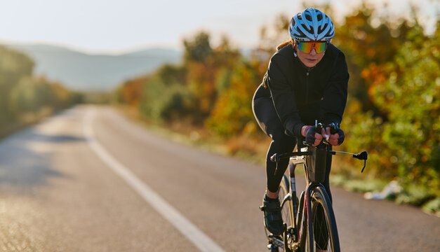 Female Road Cyclist Training on Country Road at Sunrise Wearing Helmet and Performance Sunglasses