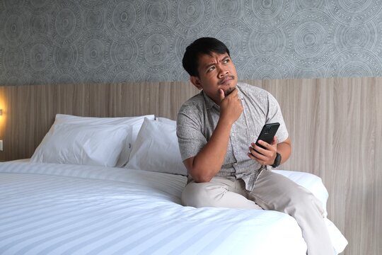 Thoughtful Asian man sitting on a bed, holding a smartphone and touching his chin while looking upward and considering an idea.