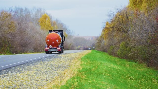 Large orange tanker truck drives down rural road. Trees show autumn colors, yellow and green grasses line path. Vehicle moves steadily toward horizon, calm and steady. Ideal for transport, logistics