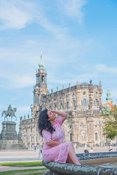 Beautiful brunette woman in pink dress sitting near fountain in Dresden, enjoying summer vacation with historic cathedral background in Saxony, Germany 