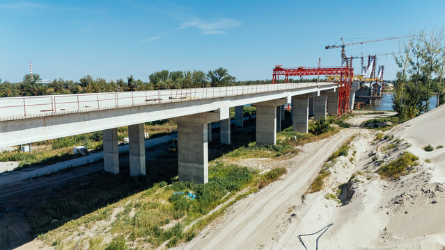 20 July 2026: Aerial view of a bridge under construction with concrete pillars, steel scaffolding, and cranes set against a clear blue sky.
