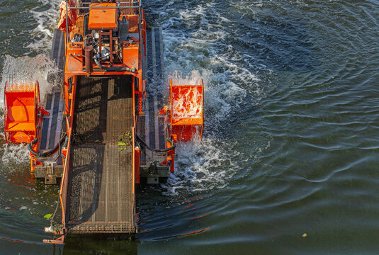 Top view of an orange industrial dredging vessel with paddle wheels churning water and collecting floating debris on a river or canal surface