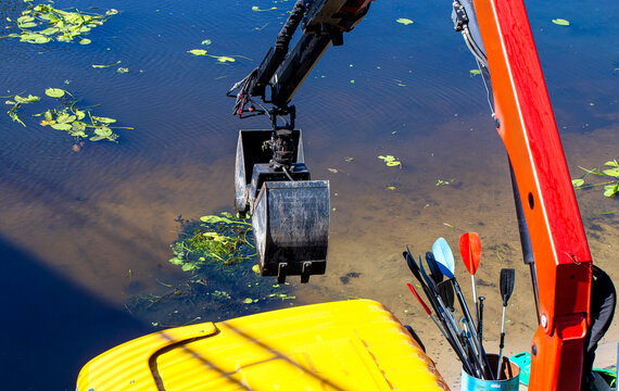 Close view of an excavator arm and bucket over calm shallow water beside paddles and a yellow boat deck at a lakeshore, suggesting shoreline maintenance or cleanup work