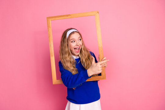 Young girl in blue sweater holds a wooden frame against pink background smiling and pointing at the frame in a playful fashion photo
