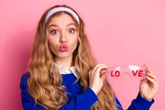 Young girl in blue sweater with pink background puckers lips while holding love glasses for playful fashion style photo