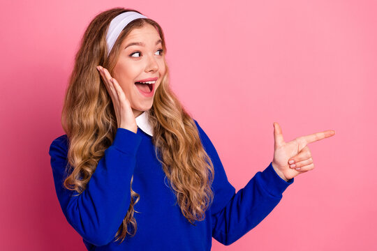 Joyful young woman in blue sweater with headband smiles and points to the right against pink background