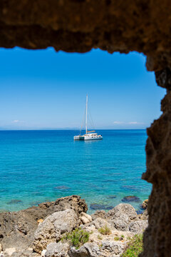 Rhodes, Greece - 16 May 2026: View of a catamaran sailing on the turquoise Aegean Sea, framed by a natural rocky archway under a clear blue sky.