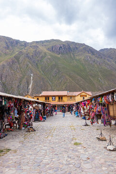 Ollantaytambo, Cusco, Peru - 05 May 2021: View of market stalls filled with colorful textiles along a cobblestone street, with dramatic mountains in the background under a cloudy sky.