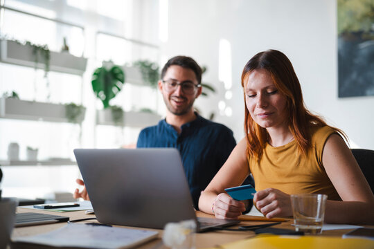 A focused woman holds a credit card while sitting at a desk with her colleague. They are shopping online or managing business finances on a laptop in an office.
