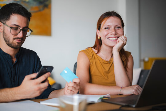 A young man holds a smartphone and credit card while working from home with a happy woman. She sits beside him and smiles at the camera as they manage personal finances together.