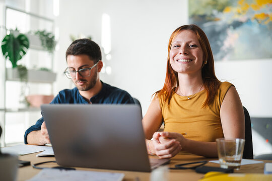 A young woman with red hair smiles at the camera while working at a desk next to her male colleague, who remains focused on his tasks in a bright modern home office.