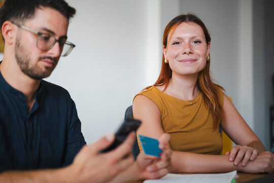 A man in glasses holds a smartphone and a payment card while sitting at a desk. A woman sits beside him and looks at the camera while they manage financial tasks.