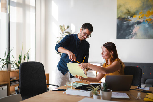 A man and a woman are working together in a bright office. They are reviewing documents and discussing a project while leaning over a wooden desk with a laptop.
