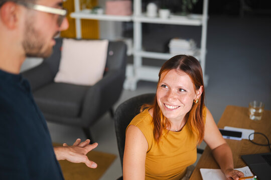 A man with glasses gestures while talking to a female colleague in a modern office space. The red-haired woman listens intently and smiles during the conversation.