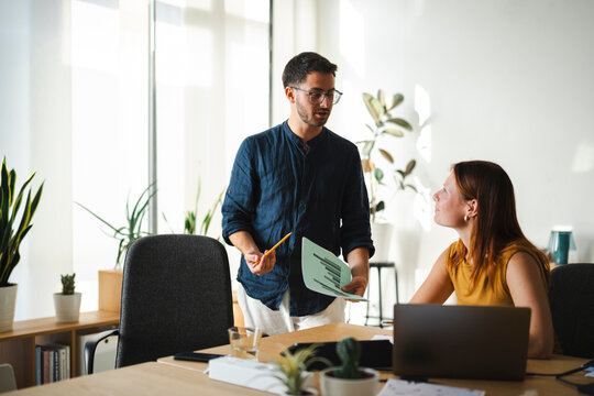 A young man wearing glasses stands and holds a business document while talking to his female colleague in a modern sunlit office. She listens while at her desk.