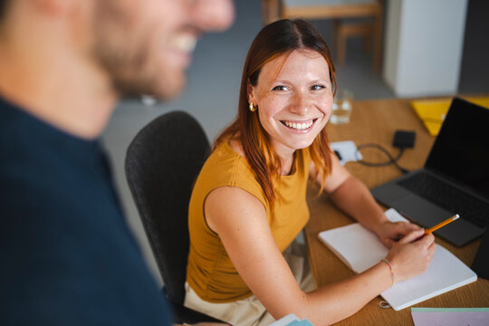 A professional woman with red hair smiles while sitting at her desk, looking up at a colleague during a friendly conversation. They are working in a bright office.