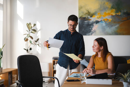 A professional man standing and a woman sitting at a desk review important documents together in a modern office. They collaborate on a creative project together.