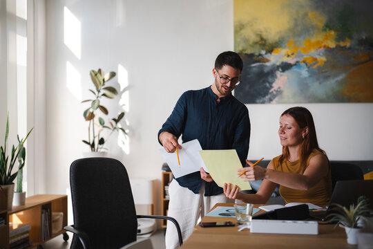 A man and a woman collaborate on a project in a sunlit modern home office. They are reviewing documents and sharing ideas together while standing at a wooden desk.