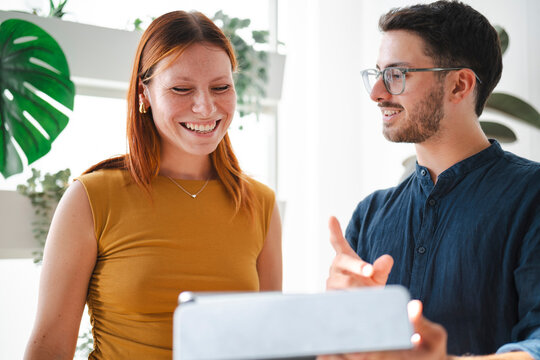 A man with glasses and a redheaded woman collaborate while looking at a digital tablet in a bright office. They discuss work projects with smiles on their faces.