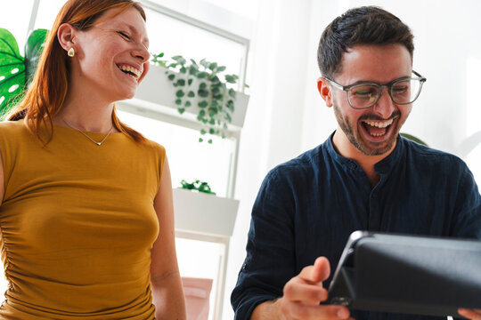A man wearing glasses and a woman with red hair laugh while looking at a tablet together in a bright office environment. They share a joyful moment while working.