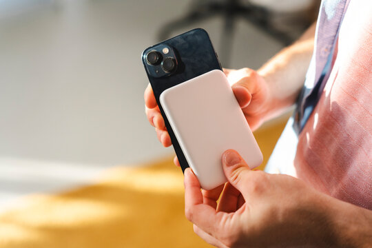 A person holds a dark smartphone with a white wireless power bank magnetically attached to the back. This portable charging accessory provides extra battery life.