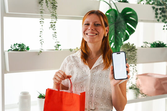 A smiling woman with red hair holds a vibrant red shopping bag and shows a blank smartphone screen. She is standing in a bright indoor garden area filled with plants.