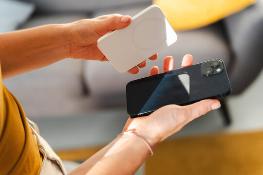 A person holds a dark smartphone and a white portable wireless power bank. They are about to attach the magnetic battery to the back of the phone to charge it.