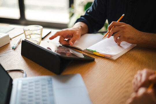 A professional collaborates on a project at a desk. He points at a digital tablet while holding a pencil and taking notes in an open notebook to organize his work.