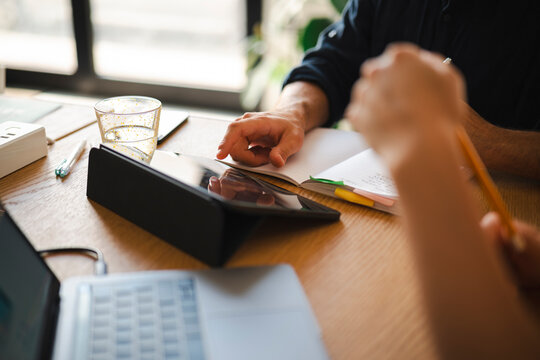Two colleagues sit at a wooden table in a sunlit office, collaborating on a project while using a tablet, laptop, and notebook to review data and share ideas together.