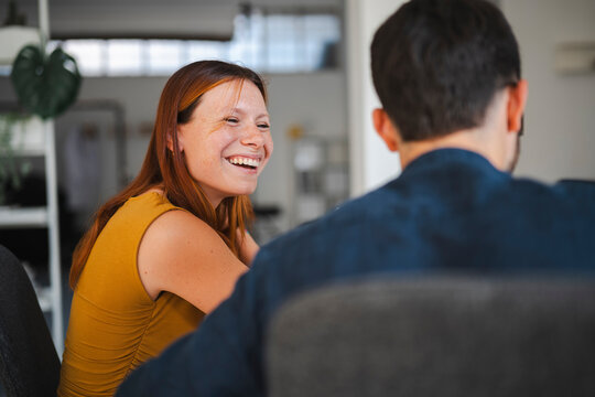 A woman with freckles and auburn hair laughs while having a lighthearted conversation with a male colleague in a bright, modern office space during the workday.
