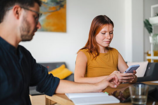 A young man and a woman work together at a desk in a home office. The woman focuses on her smartphone while the man reaches out to collaborate on a project.