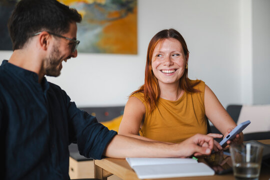A young man with glasses and a woman with auburn hair sit together at a table. They are looking at a smartphone and sharing a joyful laugh in a home setting.