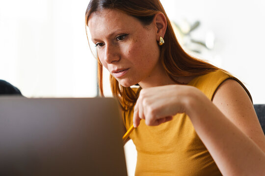 A focused woman in a yellow top works on a laptop in an office environment. She holds a pencil and concentrates deeply while reviewing data on her computer screen.