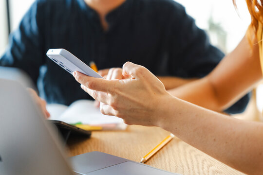A person holds a smartphone while sitting at a desk with a laptop and a pencil. A colleague works in the background during a collaborative office project session.