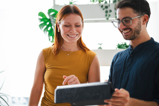 A man and woman collaborate in a modern office environment. They share a smile while reviewing data on a handheld digital tablet during a productive workday meeting.
