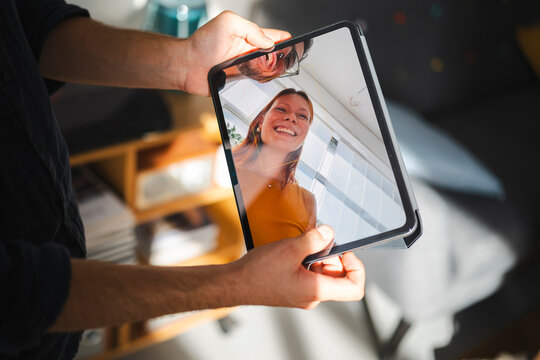 A person holds a digital tablet displaying a live video feed of a smiling woman and a man with a camera, capturing a moment of digital connection during a video call.