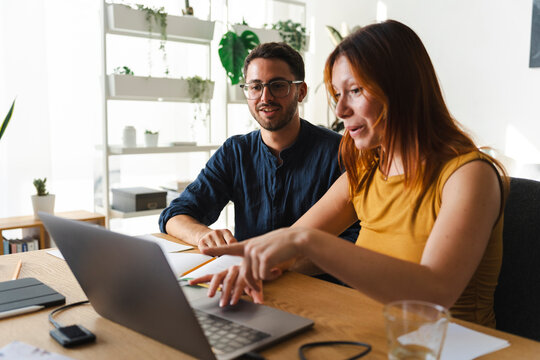 A man and a woman work together at a desk in a bright office. The woman points at the laptop screen while explaining a project to her male colleague during a meeting.