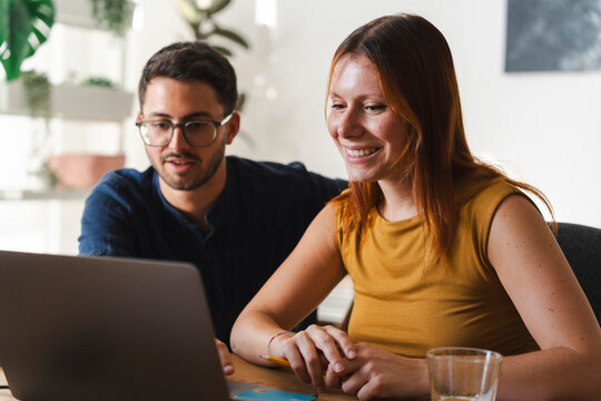 A professional man and woman collaborate on a creative project together. They sit at a wooden desk, focus on the laptop screen, and share a friendly discussion.
