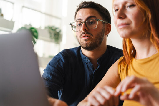 A focused man with glasses and a woman collaborate on a professional task while looking at a laptop screen together in a bright and modern open workspace environment.