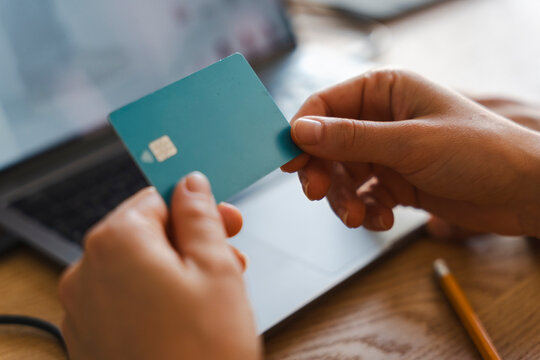 A person holds a teal debit card while sitting at a laptop computer. They are about to make an online purchase from home during a busy work session at the wooden desk.